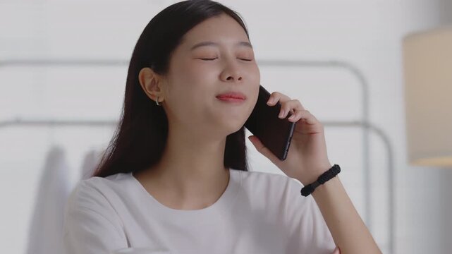 Smiling young Asian woman having a pleasant phone conversation while working at her desk in a home office, highlighting customer service, communication, and small business management.