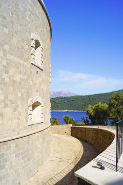 Curved outer wall of Mamula fortress with defensive gun embrasure and view of Adriatic Sea. Illustrates exclusive island resort getaway, Montenegrin scenic heritage, and maritime defense architecture.