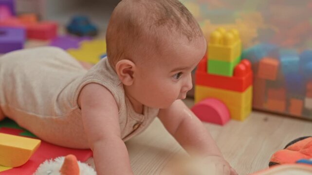 Boy reaches for colorful plastic blocks during tummy time on playroom floor with foam puzzle mat and toy tower; curious exploration.