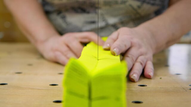 A close-up of fabric cutting. A utility knife carefully cuts through a stack of bright yellow material. The seamstress's hands hold the fabric firmly on the wooden cutting table, ensuring a smooth and