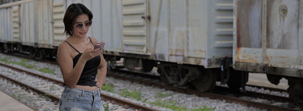 Asian woman in sunglasses, a black camisole top, and denim shorts looking down at her smartphone screen while standing on a train platform