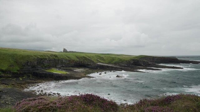 Wide coastal view of rocky cliffs, surf and purple heather at Mullaghmore Head, with Classiebawn Castle in the distance under an overcast sky.