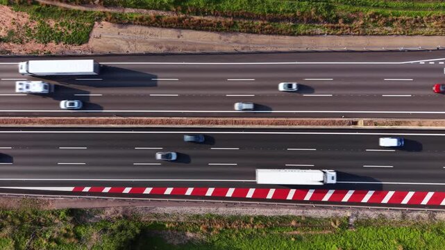 Overhead view of a multi-lane highway with two cars in motion, featuring red and white safety barriers, asphalt lanes, white dashed markings, and green roadside vegetation