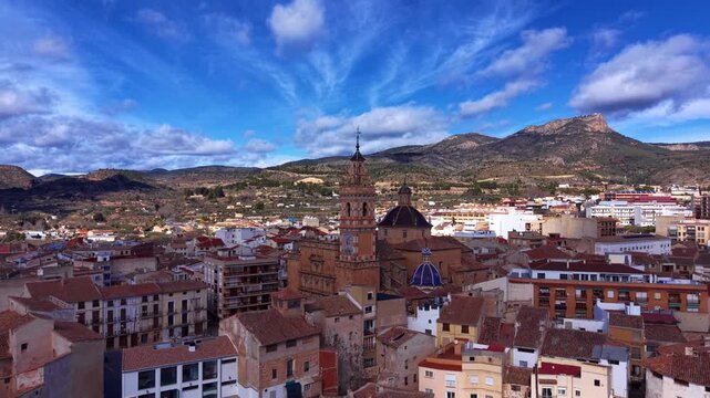 Panoramic view of Chelva, Spain, featuring a baroque church tower and blue dome rising above terracotta rooftops with mountains and blue sky beyond