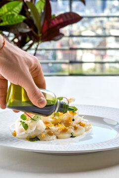 Chef dripping green herb oil onto scallop carpaccio with shaved fennel. Fine dining terrace setting with city view background.
