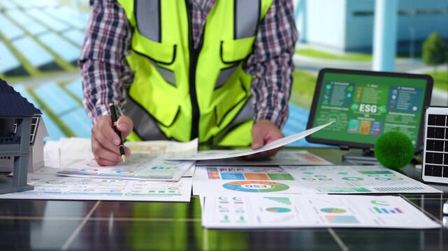 Architect in a safety vest reviews building plans and energy charts at a desk with a laptop, model house and solar panel, showing sustainable construction planning.