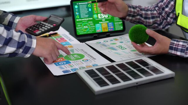 Team reviewing environmental, social and governance data with a calculator, reports, solar panel model and green microphone during a sustainability meeting.