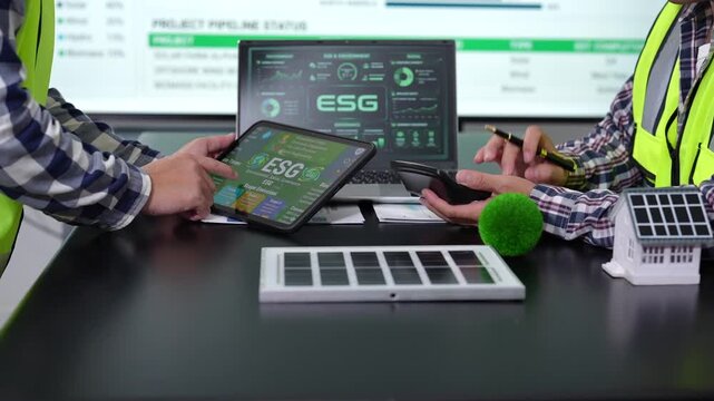 Two construction professionals review esg data on a tablet and laptop beside a model home with solar panels, discussing sustainable building plans.