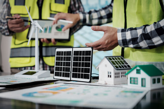 Engineers in safety vests discuss a solar energy project with panel models, wind turbine, house miniatures and charts on a planning table.