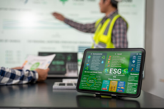 Tablet displays an esg dashboard in an office meeting while a safety vest worker presents environmental data on a board in the background.