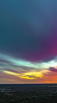 Vertical timelapse reveals iridescent pink and teal sunset clouds glowing above small village on horizon