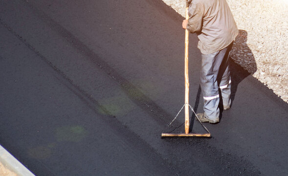 Construction worker using a raking tool to spread fresh hot asphalt on a new road surface, preparation for compaction and finishing during road repair project in bright daylight. Copy space