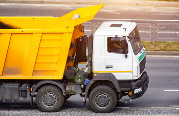 Heavy duty white dump truck with vibrant yellow dump body driving on paved city street under bright sunlight during day. Essential construction vehicle for material transport. © HENADZY