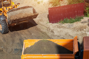 Yellow construction loader actively dumping loose soil from front bucket into large orange dump truck bed, performing earthmoving operations at outdoor construction site under bright daylight. © HENADZY