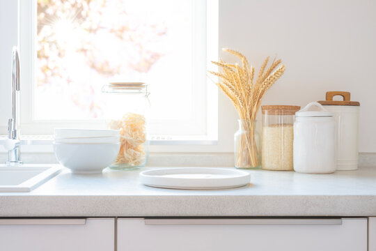 Bright kitchen counter with white plate for your design, glass jars of pasta and grains, wheat stalks by sunny window and sink