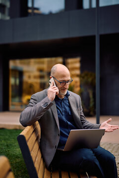 Mature businessman talking on cell phone while working on laptop outdoors.