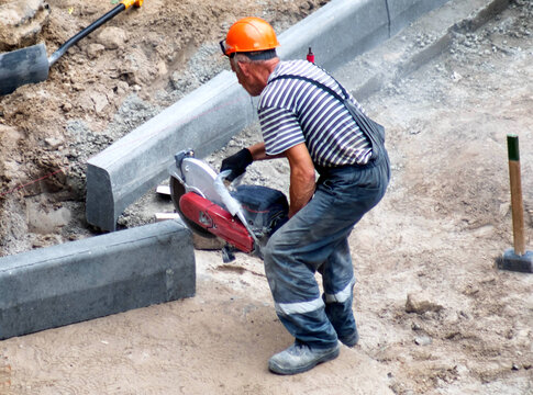 Construction worker operates power cut-off saw cutting concrete curbstone for road edge installation project. Worker wears hard hat, striped shirt, work pants, safety gloves, copy space