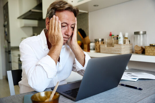 Frustrated senior man holds his head near a laptop at home