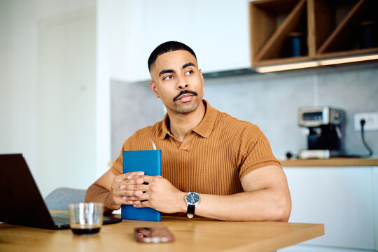 Young man contemplating an idea, holding a notebook while sitting at a kitchen table with a laptop, working remotely