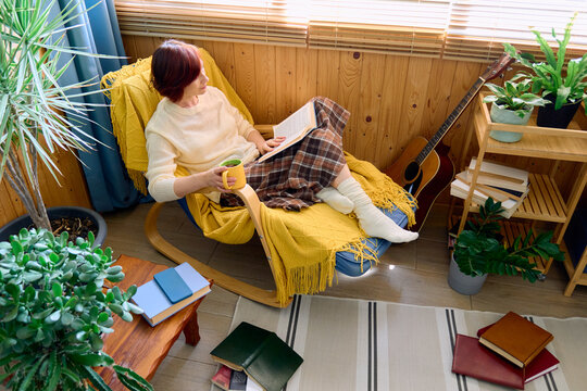 A woman reads a book and drinks tea in a cozy armchair at home