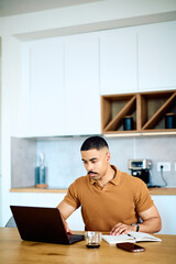 Young man using laptop and taking notes while working remotely from a modern kitchen. Focus on...