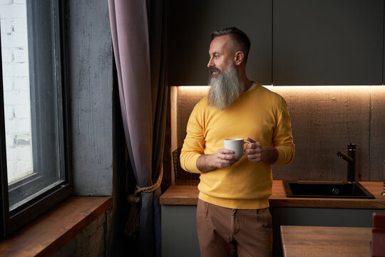 Mature man holds coffee mug and looks out window in modern kitchen