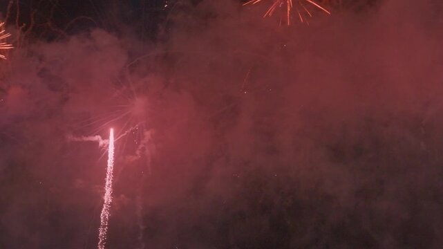 A ground-level pyrotechnic fountain shoots intense vertical sparks and bright flames behind a wooden fence along a dark roadside, filling the night air with thick smoke clouds.