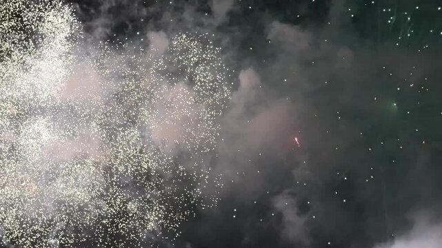 Ground-level pyrotechnics shoot brilliant spark fountains and bright flames behind a wooden fence along a dark roadside, filling the night air with thick smoke clouds.