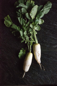 Two fresh white radishes with green leaves lie on a black stone