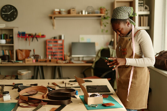 Female artisan photographs leather craft with smartphone in workshop