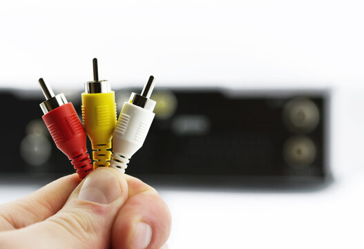 Close-up of a hand holding red, yellow, and white RCA connectors with a blurred audio video input panel in the background on a clean light backdrop