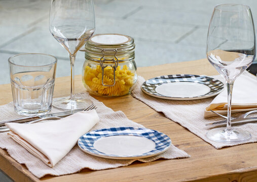 Wooden dining table arranged for two with empty wine glasses, water glass, checkered plates, folded napkins and a glass jar filled with dry fusilli pasta