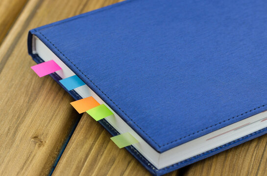 Closeup of a blue hardcover notebook with colorful sticky index tabs tucked between the pages, placed diagonally on a warm wooden tabletop