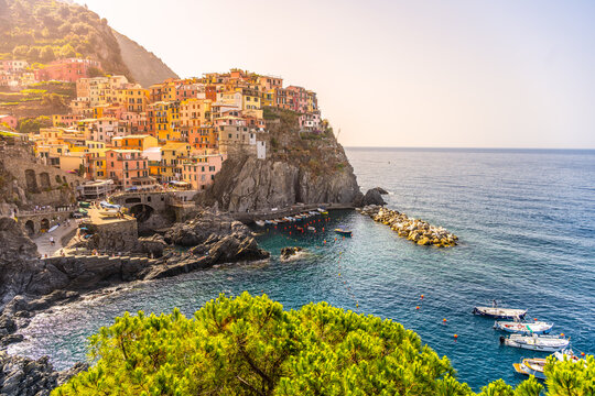 Manarola Village has colorful houses on steep cliffs above the sea. The clear water reflects the buildings and the rocky landscape. People stroll and enjoy the view during a sunny day.