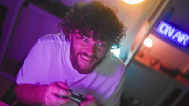 Young bearded man with curly hair holding a joystick and raising clenched fists while cheering in a streaming studio lit by neon signs; triumph.