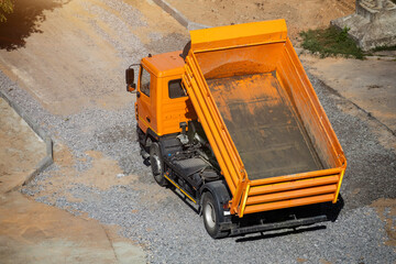 Orange dump truck with elevated bed on gravel road at construction site, ready to unload bulk materials like crushed stone or sand, sun shining on vehicle surface, copy space © HENADZY