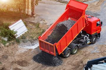 Red dump truck actively unloads crushed stone material onto construction ground. High angle view of heavy vehicle engaged in material delivery for building development project under bright sunlight. © HENADZY