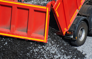 Red dump truck tailgate open, unloading bulk dark crushed stone onto pile of mixed aggregate. Construction heavy equipment operation, gravel material delivery at building site. © HENADZY