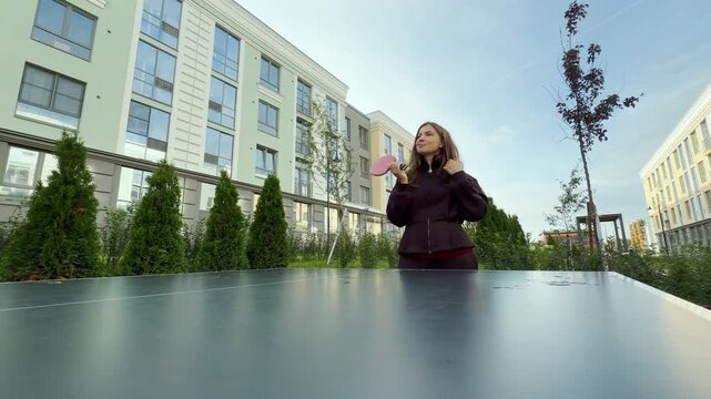 A serene woman holds a pink ping pong paddle with a confident smile. She rests between table tennis games at an outdoor table. Modern residential buildings line the bright courtyard.