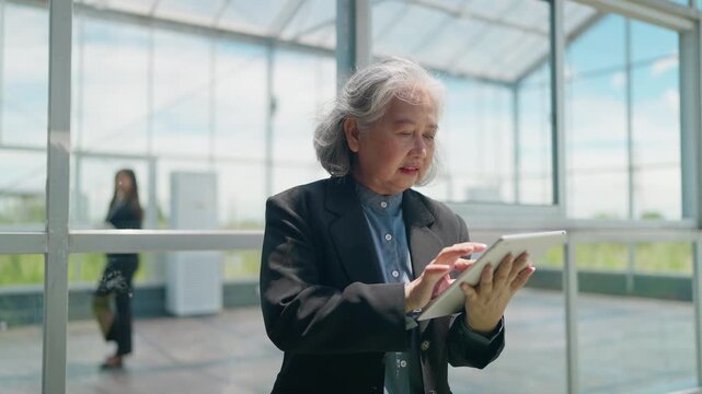 Senior business smiles while using tablet inside greenhouse. She stands near large glass walls. Bright daylight shows green plants outside
