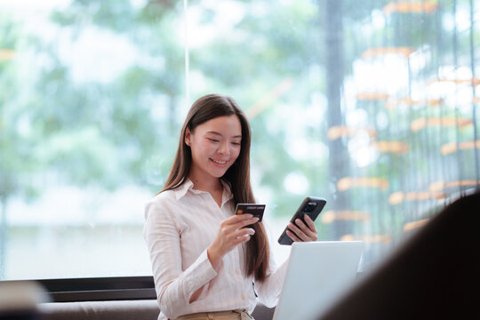 Young Asian woman using a smartphone and credit card beside a laptop, representing online shopping, digital payment, e-commerce, banking, and modern lifestyle.