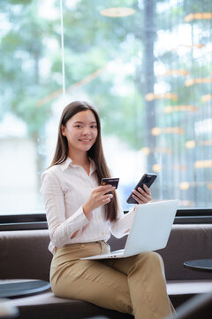 Young Asian woman using a smartphone and credit card beside a laptop, representing online shopping, digital payment, e-commerce, banking, and modern lifestyle.