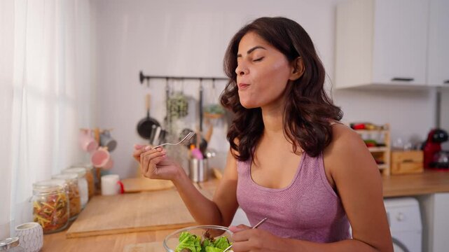 Attractive Indian woman eating healthy green salad in kitchen at home. 