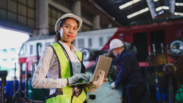 Female railway worker holding tablet in safety vest inside locomotive maintenance workshop