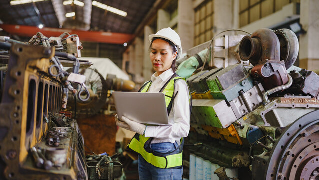 Female engineer in safety helmet and vest using laptop between aircraft engine parts inside industrial maintenance workshop, focused on mechanical inspection and repair work