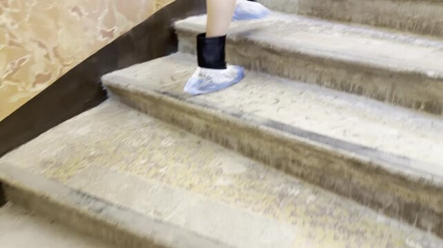 A person wearing shoe covers walks steadily up stone stairs. The individual ascends with quiet purpose inside an old building. The surroundings evoke a sense of calm discovery and mystery.
