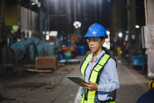Female industrial engineer wearing blue helmet and safety vest holds tablet inside manufacturing facility, representing workplace inspection, engineering, and occupational safety in heavy industry