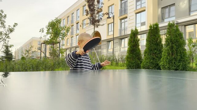 An excited young boy swings a table tennis paddle with intense joy. He plays actively behind an outdoor ping pong table. Modern apartment buildings and green trees frame the cheerful courtyard.