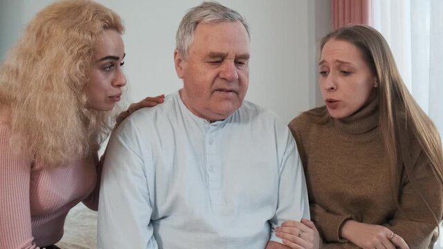 Loving daughters comforting sad elderly father, hugs him while sitting on the bed. Women caring older man at home. Family support, communication, relationships and mental health