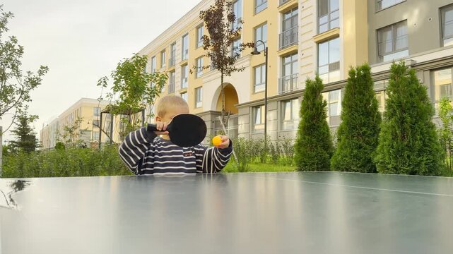 A playful young boy covers his face with a black ping pong paddle while holding a yellow ball. He enjoys a silly game at an outdoor table.